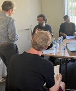 A group of people sitting at a table, one of them is talking to someone who is leaning against the wall of the room