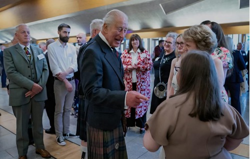 His Majesty, King Charles, smiles and points towards a young woman with her back to the camera, Megan. King Charles wears modern Highland dress and Megan is wearing a pale mushroom top and has mid-length fair hair.