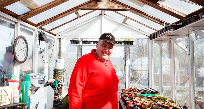 A man standing in his large greenhouse. There is plants and gardening equipment around him. He is wearing a red sweatshirt and a black NY branded baseball cap. He is smiling and not looking directly at the camera.