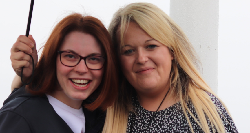 Two women standing close together. The woman on the right has her arm around the other woman and she is holding an umbrella. In the background you can see the sea but only faintly as it's wet day.